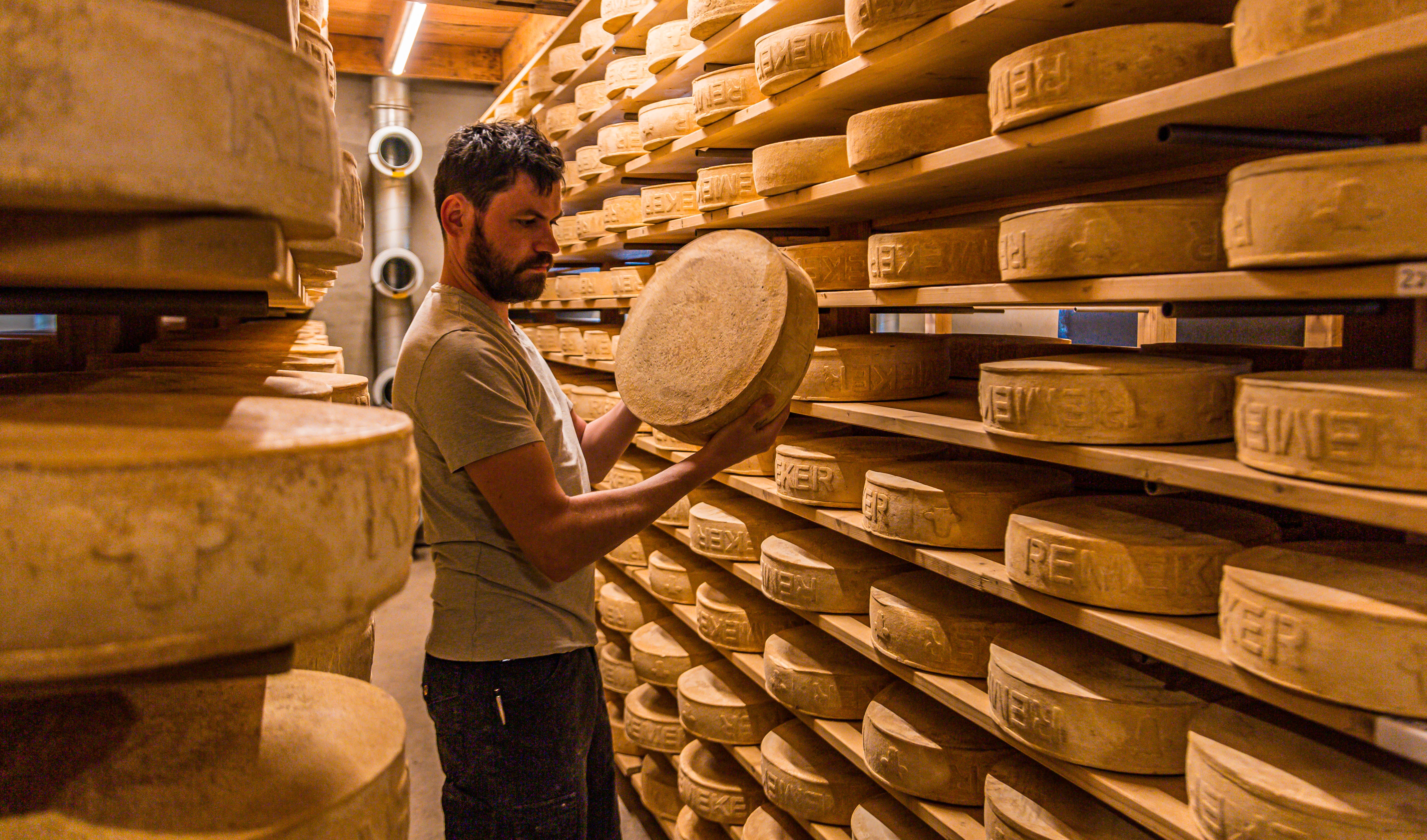 Een kaasboer staat in een kaasopslag en laat een kaas zien.